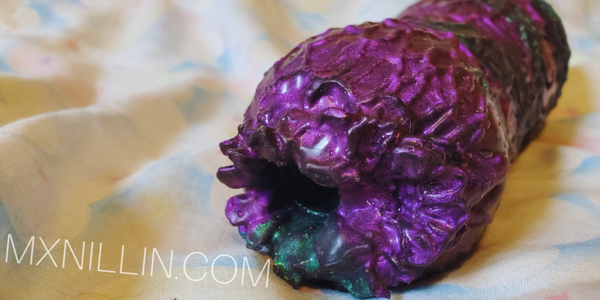A close-up of a textured, metallic purple and green silicone object resting on a light-colored fabric surface. A white watermark reading 'MXNILLIN.