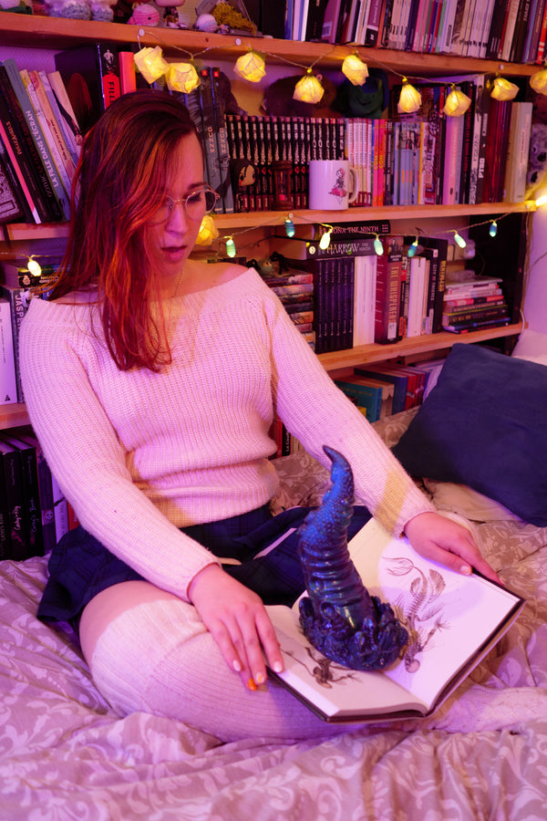 A woman with glasses sits on a bed in a cozy, book-filled room, holding an open book with a dark horned creature illustration on the page. She is wearing a light pink ribbed sweater and dark shorts, with warm fairy lights and bookshelves in the background.
