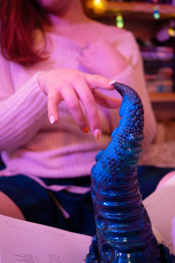 A person in a ribbed knit sweater gently touches a glossy, textured blue object in the foreground, with a softly blurred indoor background and warm string lights.