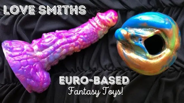 Werbefoto eines lilafarbenen Fantasy-Dildos und eines metallisch blau-goldenen Strokers auf schwarzem Stoff mit dem Text „Love Smiths – Euro-Based Fantasy Toys“.