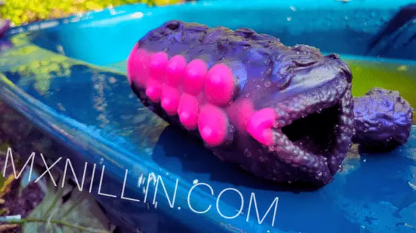 Außenaufnahme eines lilafarbenen Fantasy-Strokers mit leuchtend pinken Saugnapfstrukturen auf einer blauen Oberfläche, mit dem Wasserzeichen „MXNILLIN.COM“.