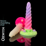 Godemiché fantasy Orwyn Unicorn Shape-Shifting Druid en silicone, rose pastel et lavande avec base sculptée verte, présenté sur fond noir brillant réfléchissant avec branding du nom du produit.