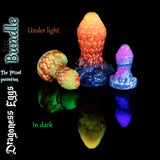 Dragoness Eggs fantasy butt plugs under light and glow-in-the-dark effect, showing textured dragon scale silicone anal plugs in vibrant orange, green marble and purple custom blends.