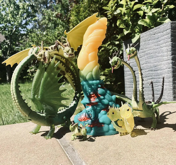 Verspielte Außenszene mit mehreren grünen Drachen-Actionfiguren rund um einen großen zweifarbigen gelb-blauen, strukturierten Fantasy-Dildo auf einer Terrasse.