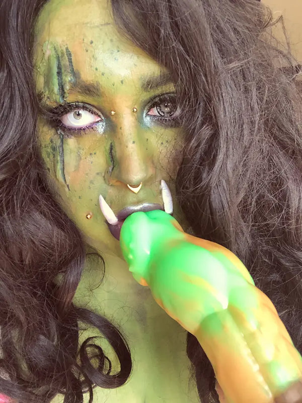 Fantasy-Cosplay in Nahaufnahme mit einer Person in grünem Ork-Make-up und Hauern, die intensiv in die Kamera blickt und mit einem leuchtend grün-orange gestreiften Fantasy-Lustprodukt interagiert.