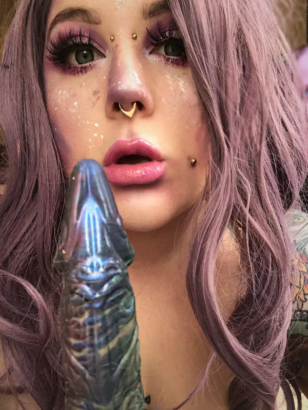 Fantasy-Cosplay in Nahaufnahme mit einer Person in aufwendigem lilafarbenem Elfen-Make-up und passender Frisur, die ein schimmerndes dunkelblaues, strukturiertes Silikontoy nahe an ihrem Mund hält.