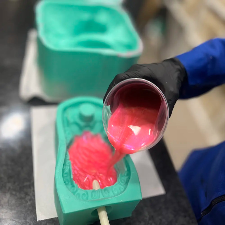 Nahaufnahme des Herstellungsprozesses: Eine Person in blauem Overall und schwarzen Handschuhen gießt in einer Laborumgebung schimmerndes pinkes Flüssigsilikon in eine türkisfarbene individuelle Form für ein Lustprodukt.